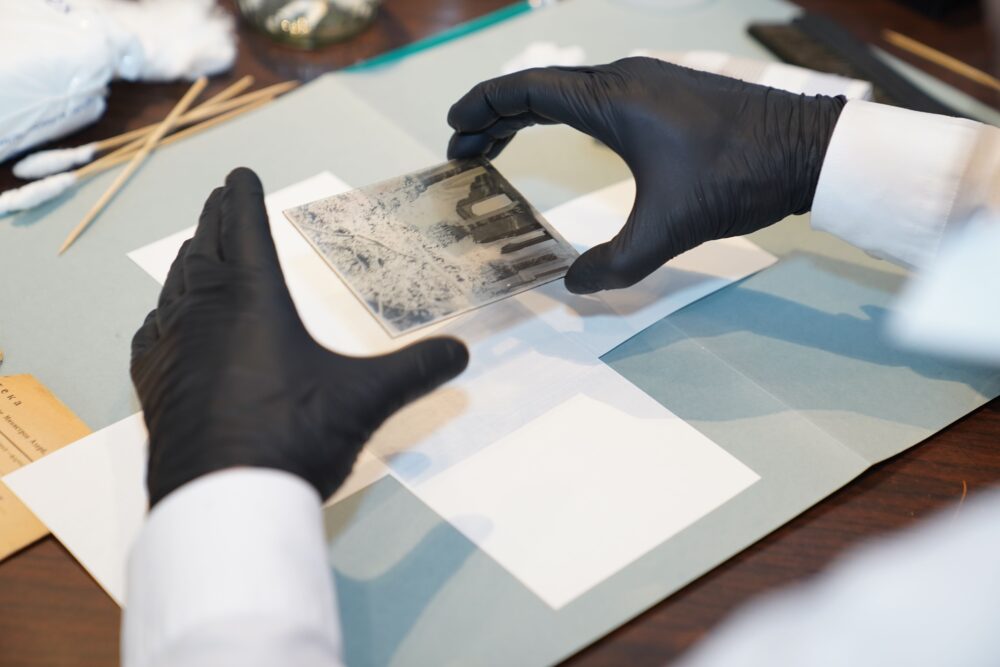 Close-up of a historic glass plate photographic negative held against a light table with white gloves, dust and...