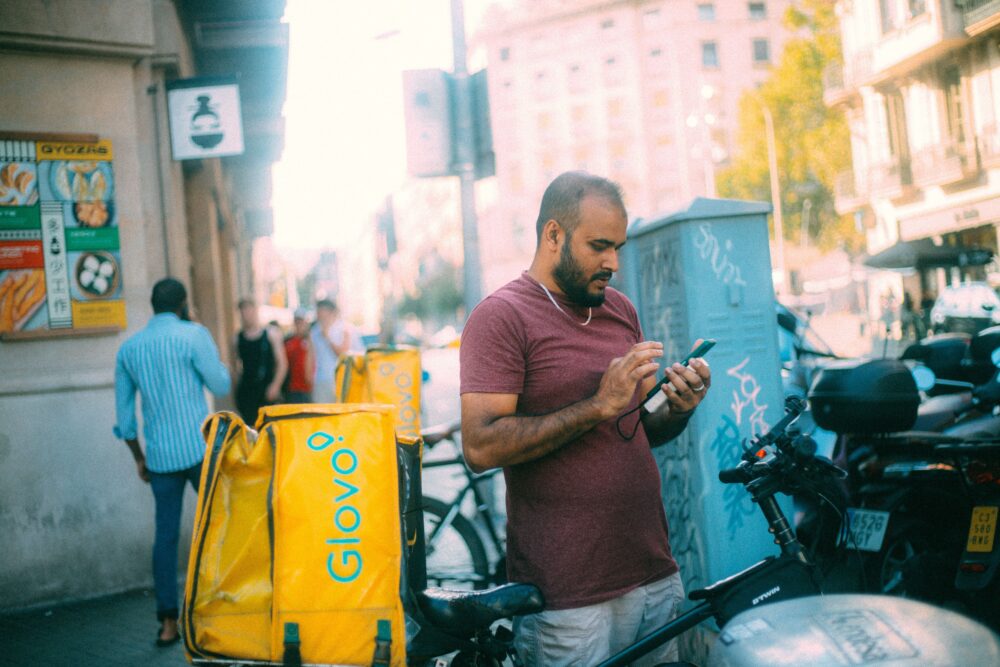 Food delivery courier on a motorbike checking a smartphone app at a traffic light in a dense São Paulo street,...